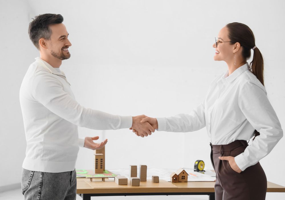 The Modern Art of Negotiation; Two people shaking hands across a table with architectural models and blueprints.