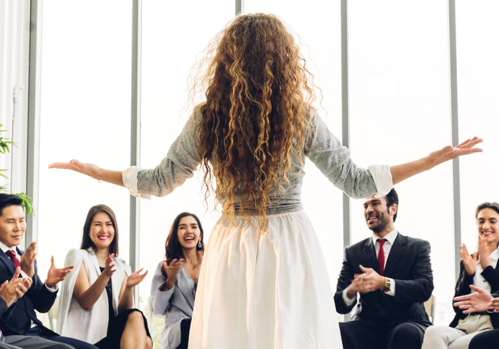 workshop: A woman with long, curly hair stands with her arms outstretched, facing a group of people who are clapping and smiling.