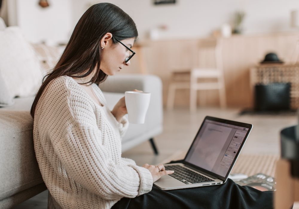 content funnel: A person wearing glasses and a cream-colored sweater sits on the floor holding a white mug and typing on a laptop.