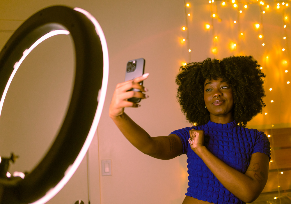 creator: A person is taking a selfie with a phone in front of a ring light, with fairy lights in the background.