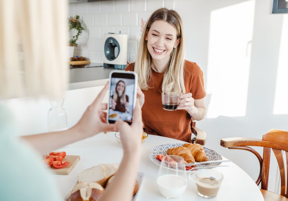 User Generated Content: A person holds up a smartphone to take a picture of another person holding a cup of coffee and smiling at a table with breakfast items.