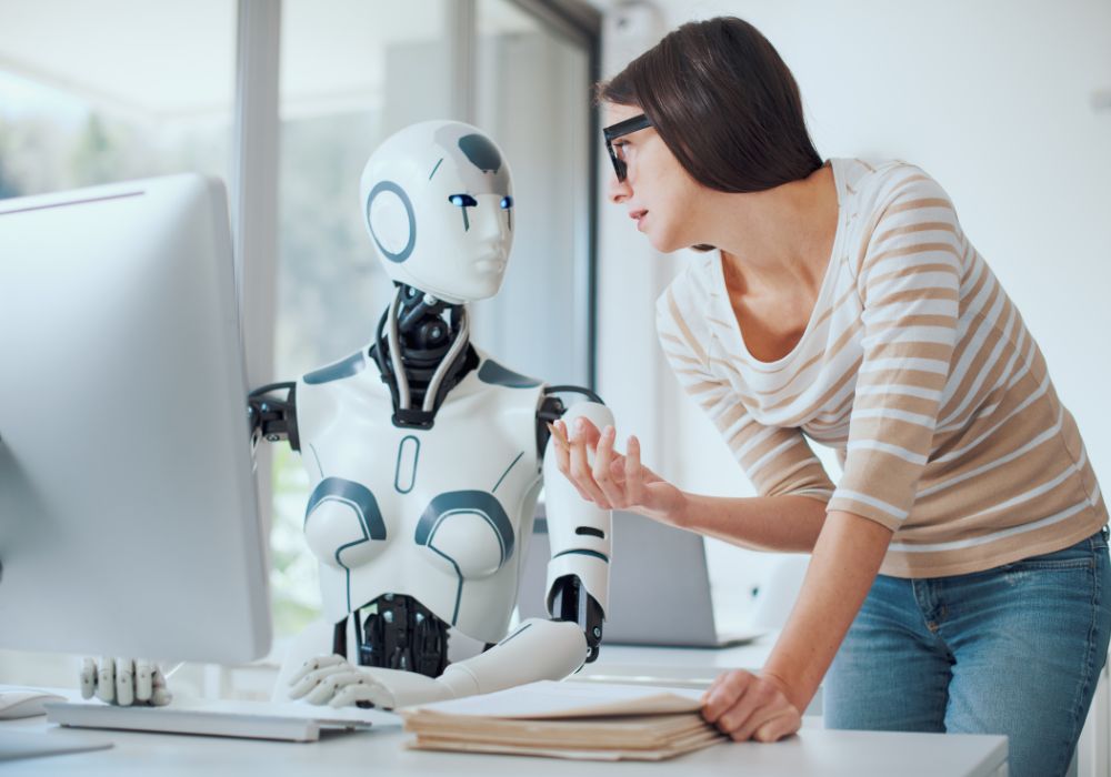 Cold email marketing: A person wearing glasses and a striped shirt talks to a humanoid robot sitting at a desk in front of a computer.